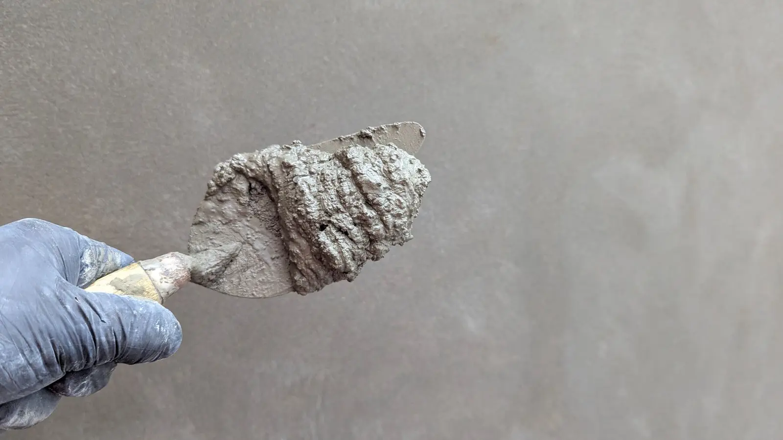 Une main gantée tient une truelle chargée de mortier gris, devant un mur enduit.